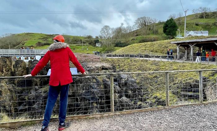 Person standing in foreground looking across a chasm in the ground towards a bridge where a small group of people stand.  On the right hand side of th